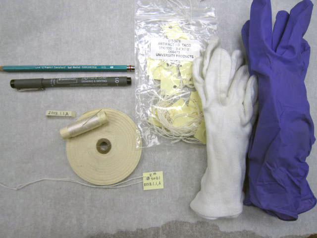 Curatorial supplies laid out on a table. Includes a blue pencil, black pen, needle and thread, cloth tags, paper tags, cotton gloves, and plastic gloves.